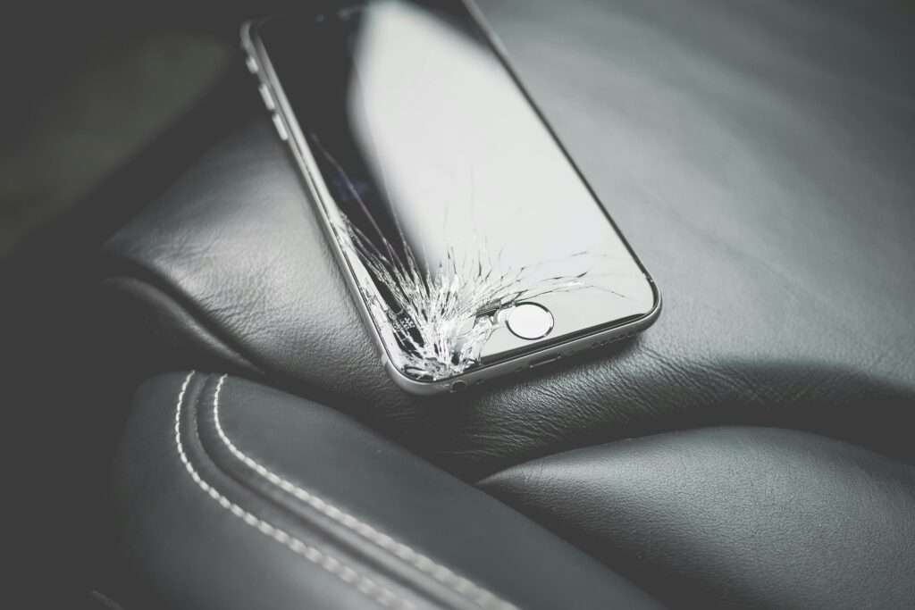 Close-up of a broken smartphone with a cracked screen on a leather car seat.